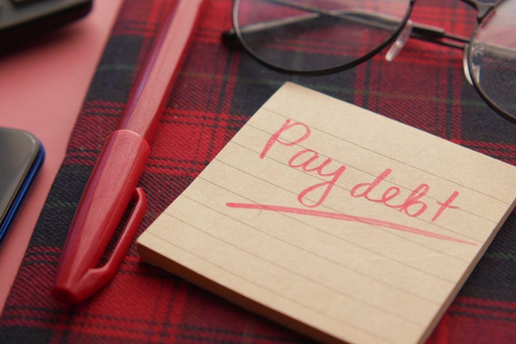 Close-up of a note reading Pay debt next to a red pen on a plaid fabric, emphasizing financial reminders.